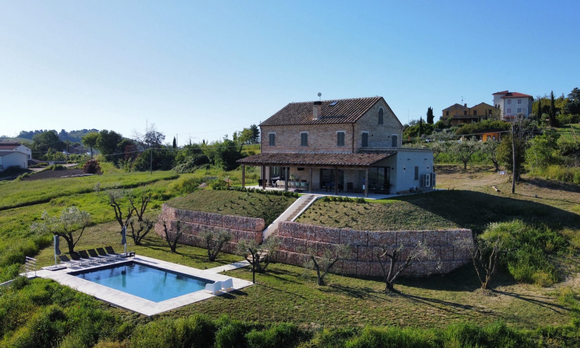 Vakantievilla Casa Camini in Le Marche, gebouwd in traditionele baksteenstijl met overdekt terras en uitzicht op het privézwembad en olijfbomen.
